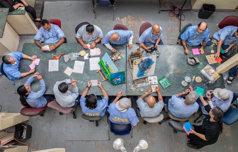 A group of people sitting around a table doing art at a prison.