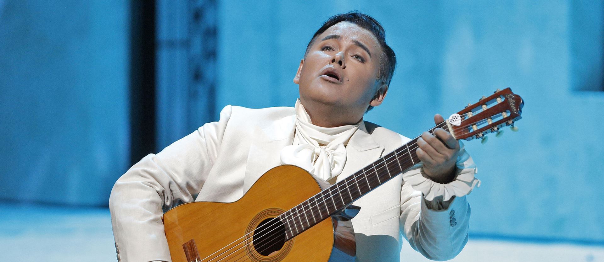 Person sitting on edge of stage in a white tux singing and playing guitar