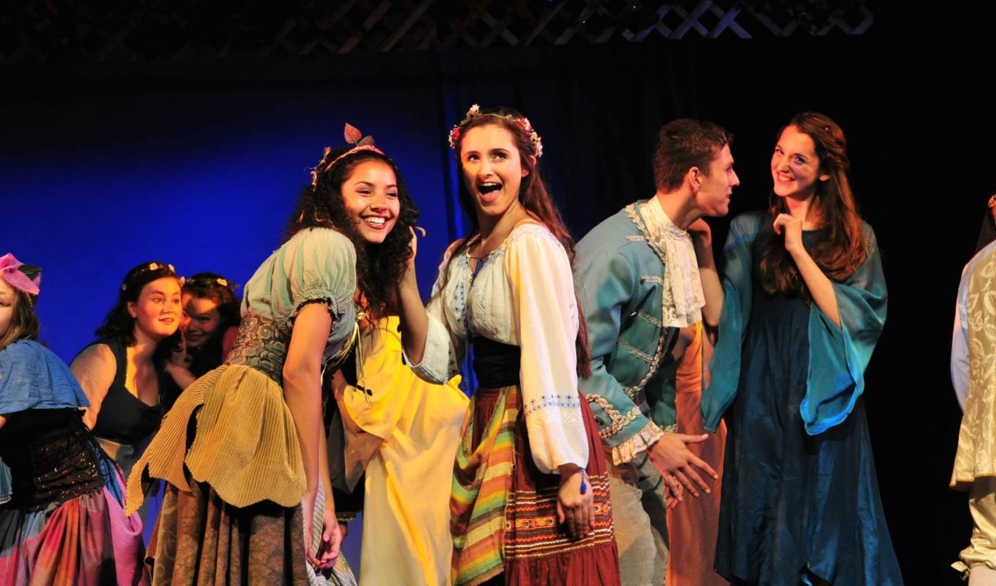 two girls singing with other castmates behind them appearing to be whispering