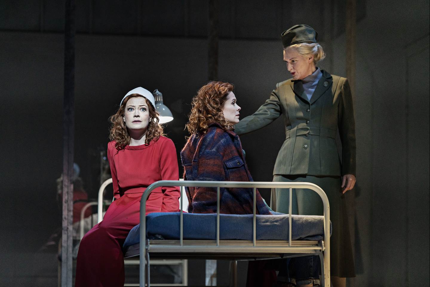 A handmaid sitting on the edge of a bed, with a scared look, while another woman is sitting on the other side of the bed being spoken to by a woman dressed in military style clothing.