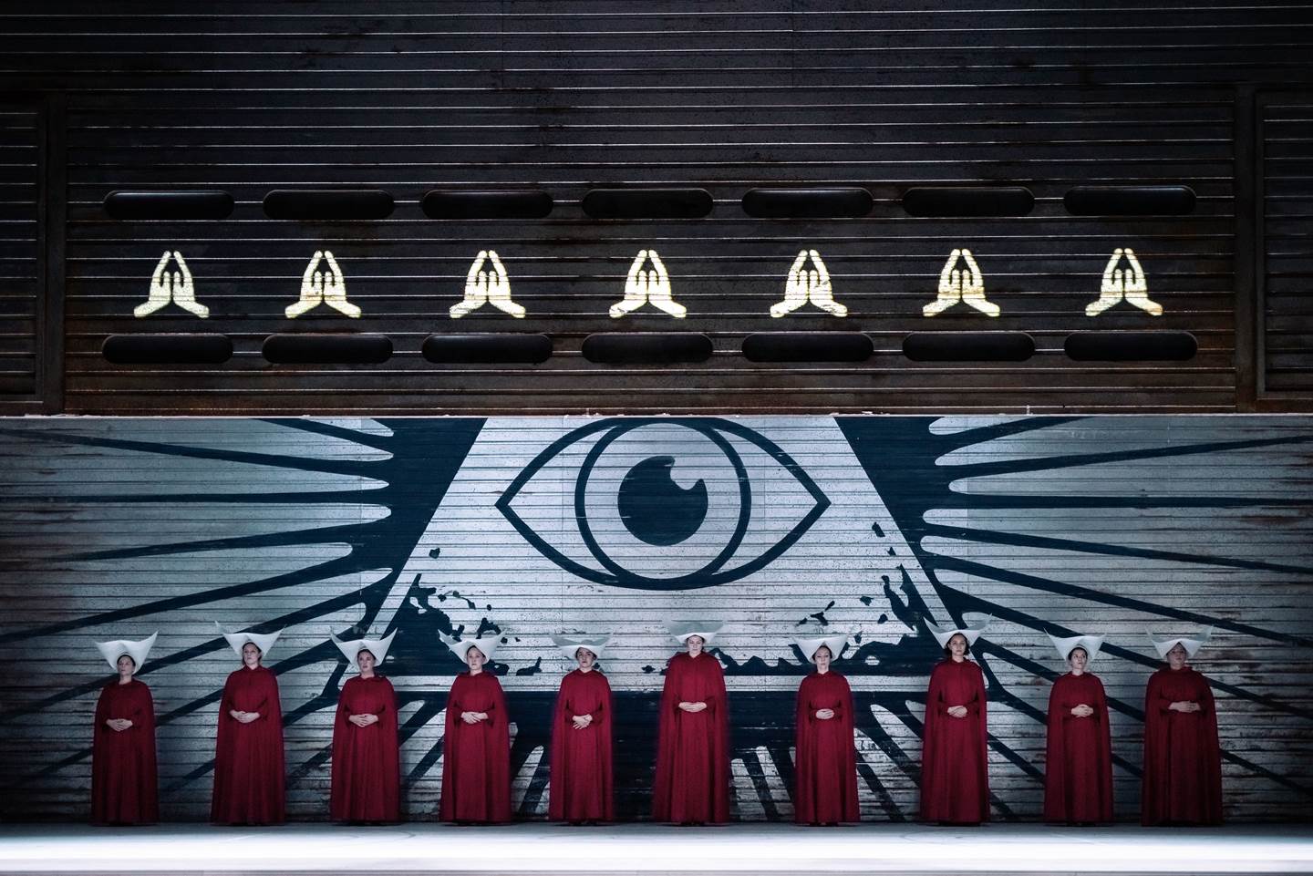 A group of handmaids in a row against a wall with a large eye in the center of a pyramid painted on it.