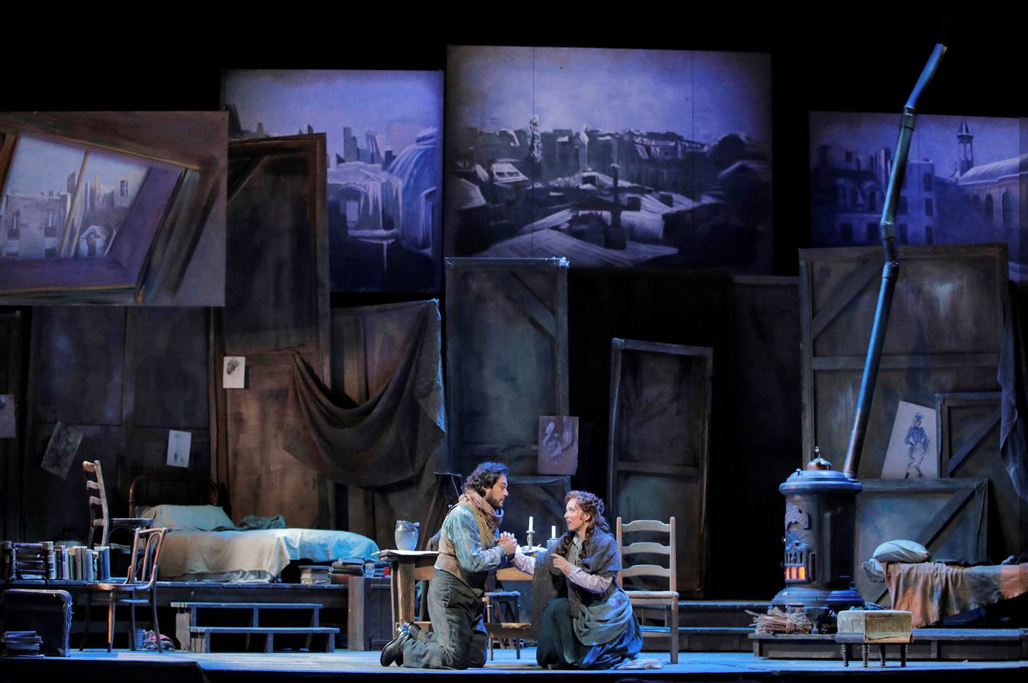 Two cast members from La Boheme kneeling on the floor of a rundown studio holding hands