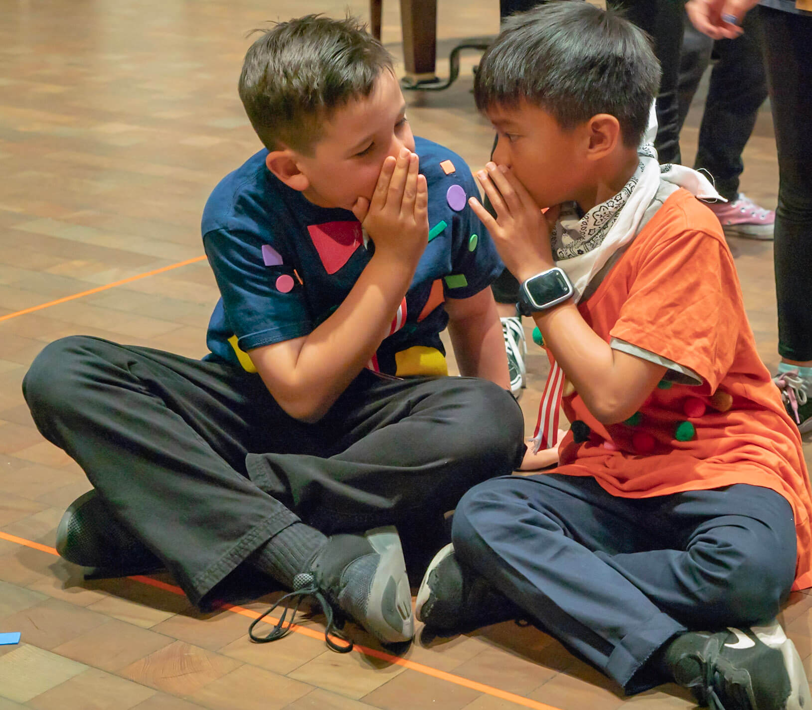 Two children sitting on the floor whispering to each other