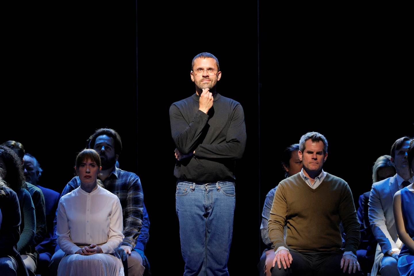 man contemplating with hand under chin in spotlight with multiple people sitting behind him