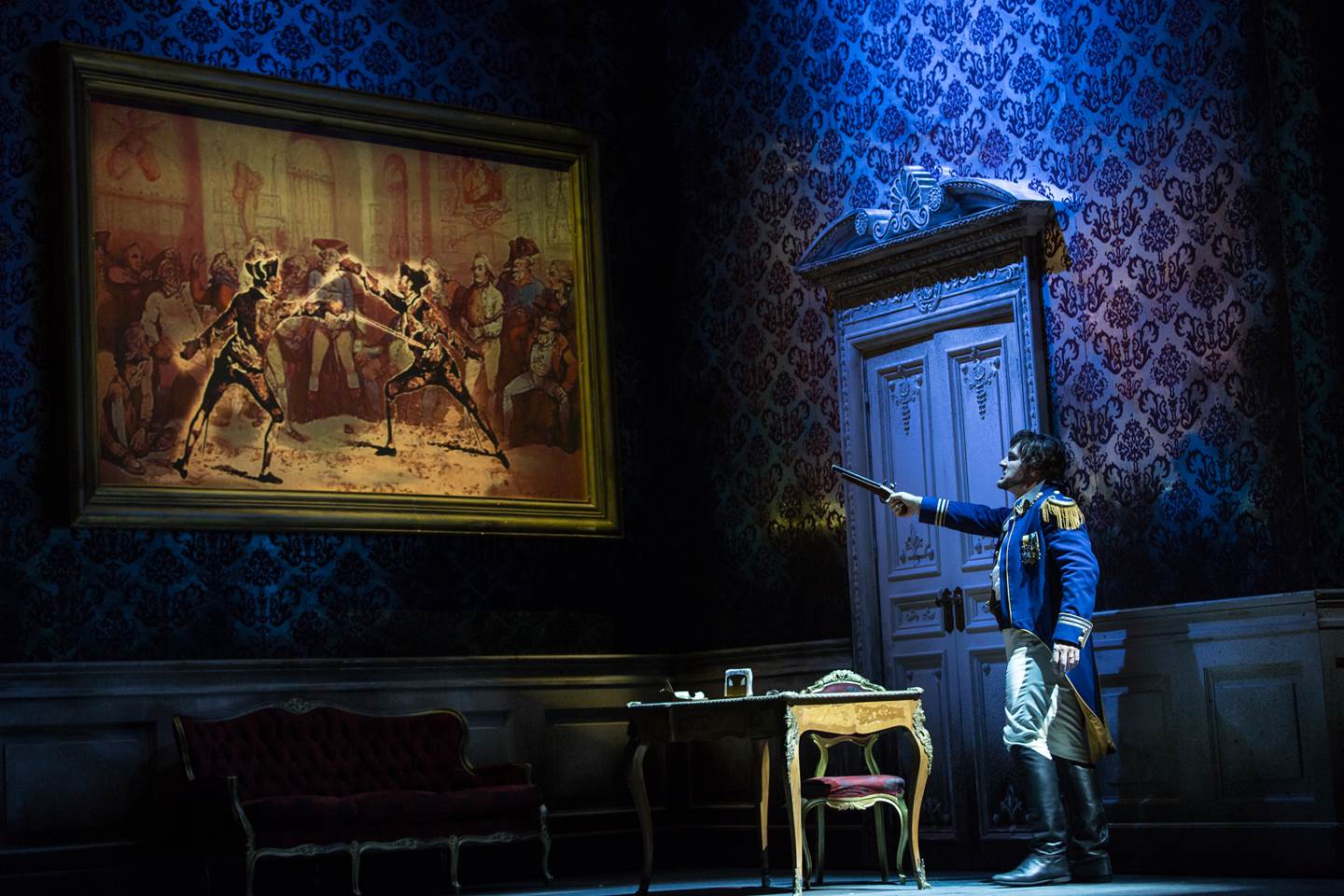 A male cast member in uniform points his gun at a painting on the wall of two men sword fighting.