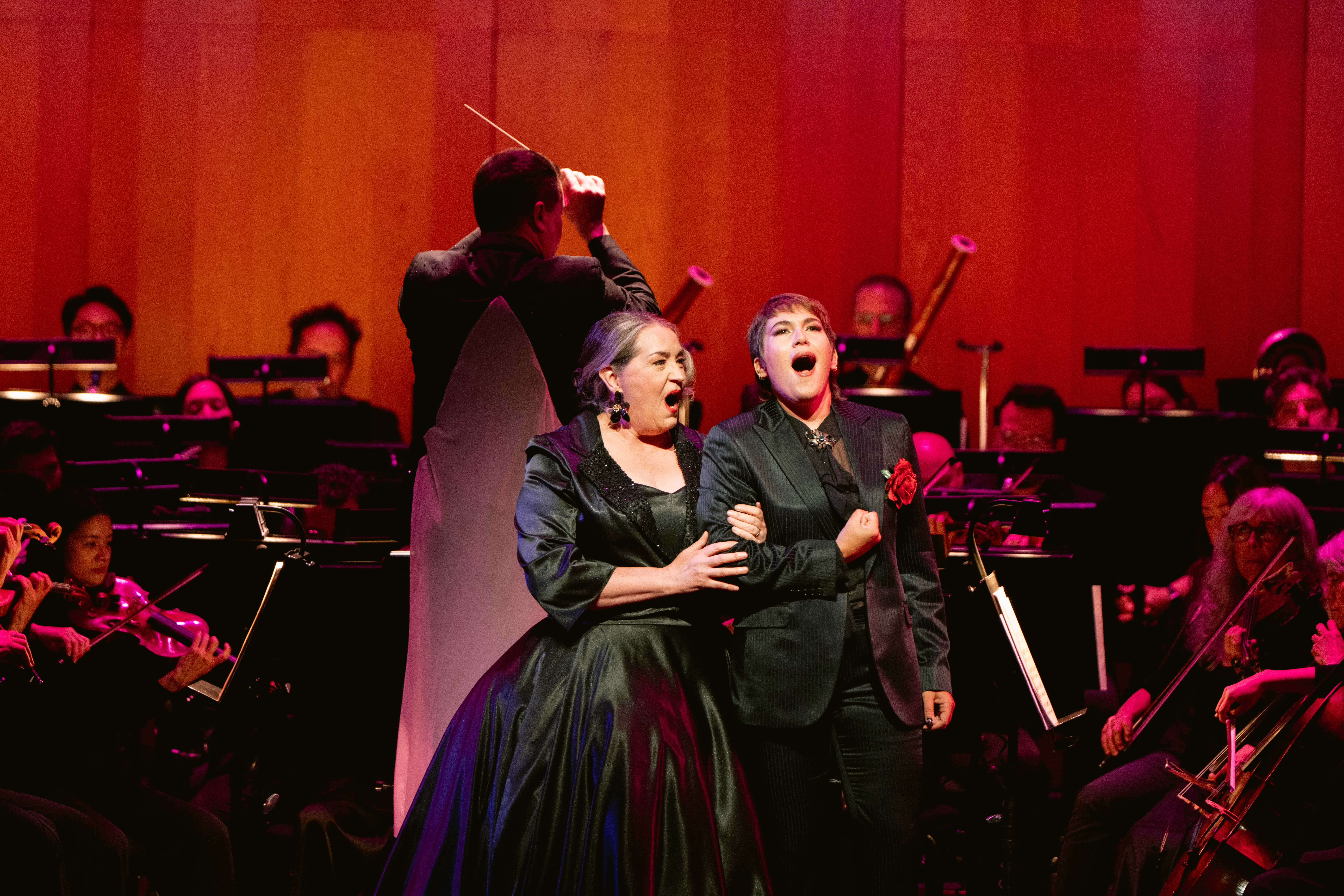Two singers wearing black singing in front of the San Franciso Opera Orchestra under red lighting.
