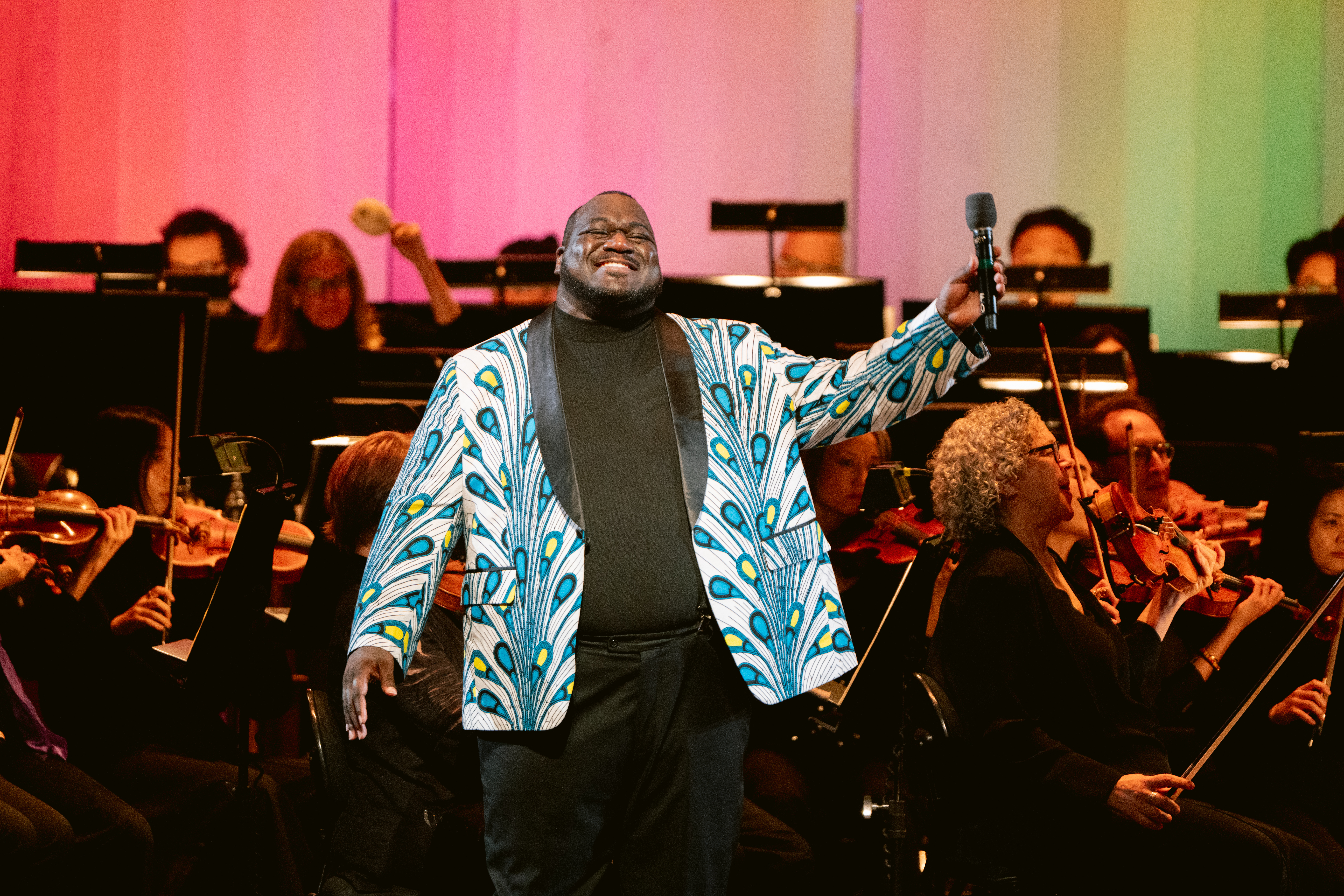 Pride concert performer standing in front of the San Francisco Opera Orchestra beneath rainbow lighting.