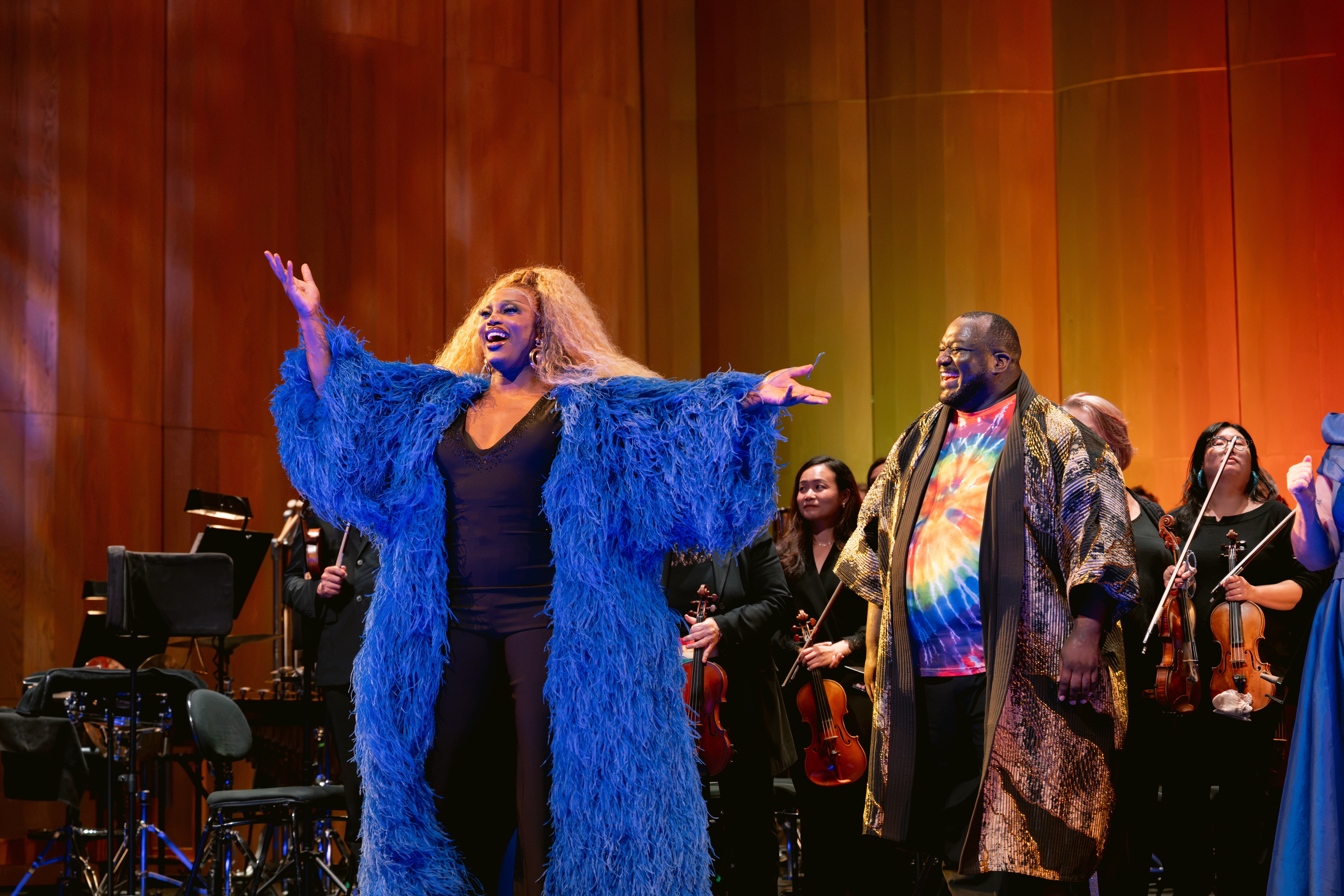Performers at the 2026 Pride Concert sing with the San Francisco Opera Orchestra, one in a blue feathered coat and one in a rainbow shirt.